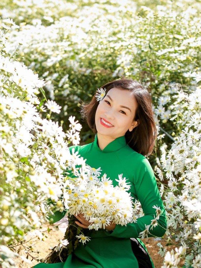 Chuyên gia Đặng Thị Hồng Phúc mặc áo dài xanh lá, tóc ngắn, cài một bông cúc hoạ mi trên tóc, ôm bó cúc hoạ mi trắng và mỉm cười giữa vườn hoa.