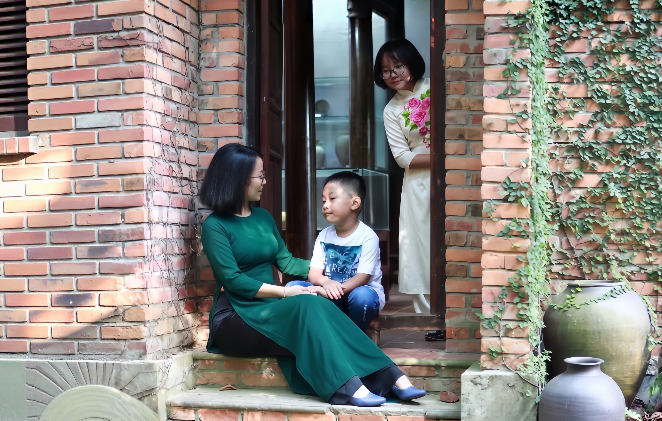 Mẹ mặc áo dài xanh ngồi trên bậc thềm gạch, nghiêng người nắm tay con trai đang ngồi cạnh; phía sau, một người phụ nữ mặc áo dài trắng đứng tựa khung cửa nhìn hai mẹ con.